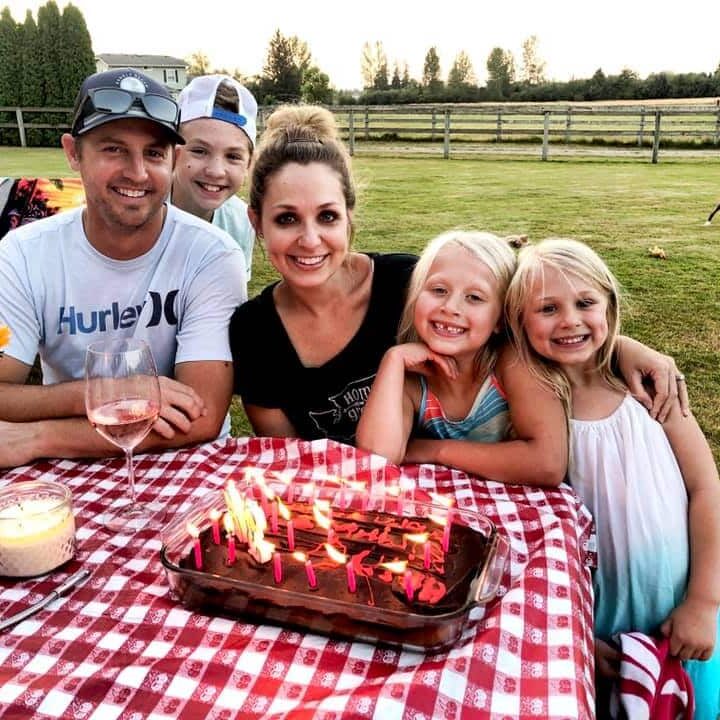 A family of four sits at an outdoor table with a red checked cloth, decorated with flowers, a candle, and a lit birthday cake. The setting shines just like surfaces treated with protective coatings in Auburn WA—perfect for special moments.