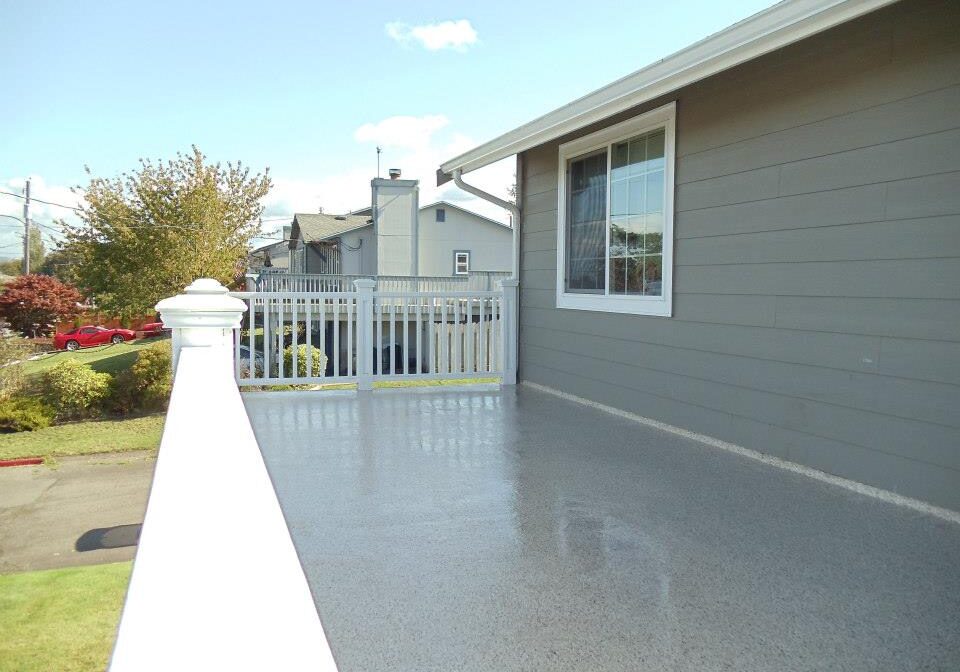 A clean, gray balcony with a white railing—finished with protective coatings Auburn WA—is attached to a gray house; a window and neighboring houses are visible in the background.