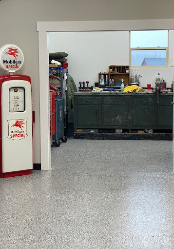 A room with a retro Mobilgas pump, a wooden display cabinet, residential and commercial flooring Auburn WA, a wall-mounted TV, and a view into a workshop area with tool cabinets and various items.