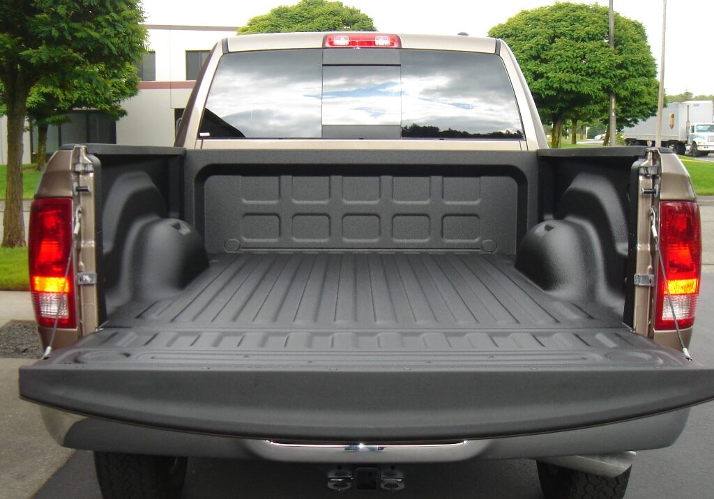 Open pickup truck bed with black spray-on truck bed liner, tailgate down, parked outdoors near trees and buildings in Auburn, viewed from the rear.
