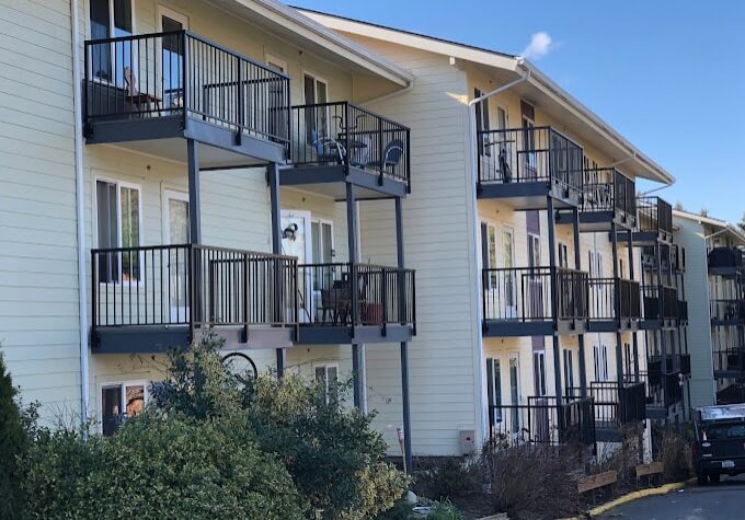 Three-story apartment building with multiple balconies featuring deck and patio waterproofing Auburn WA, plus outdoor chairs and railings. Bushes and plants line the ground floor, and a car is parked at the far end.