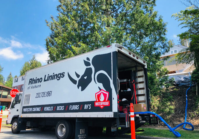 A white Rhino Linings of Auburn truck, specializing in residential and commercial flooring in Auburn WA, is parked on a street beside trees and houses, with equipment and blue hoses extending from its open rear door.