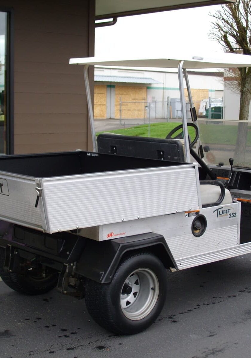 A utility golf cart with a flatbed cargo area is parked outside a building, next to a tree and an industrial area—perfect for transporting supplies or checking out Rhino Linings of Auburn FAQs on durability.