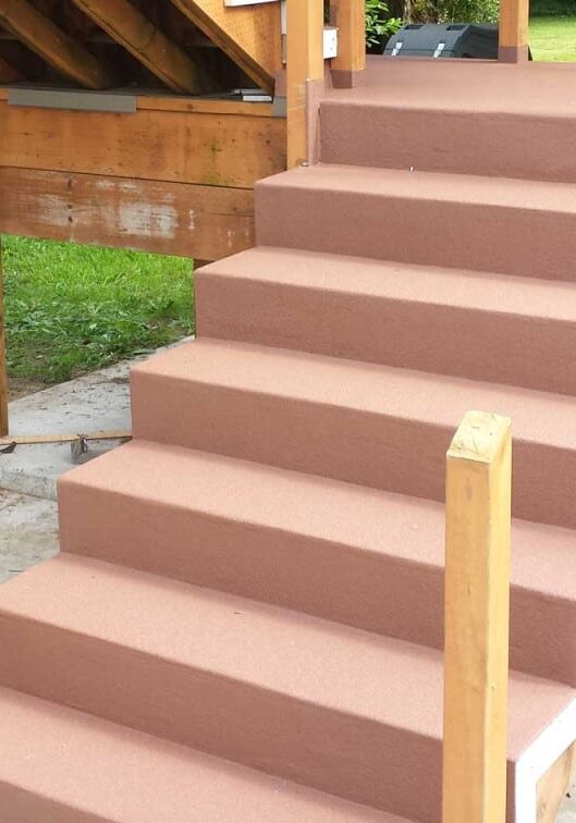 Outdoor stairs with brown steps lead up to a wooden deck treated with protective coatings. The area features support posts, potted plants, and a green lawn in the background—ideal for homes in Western Washington.