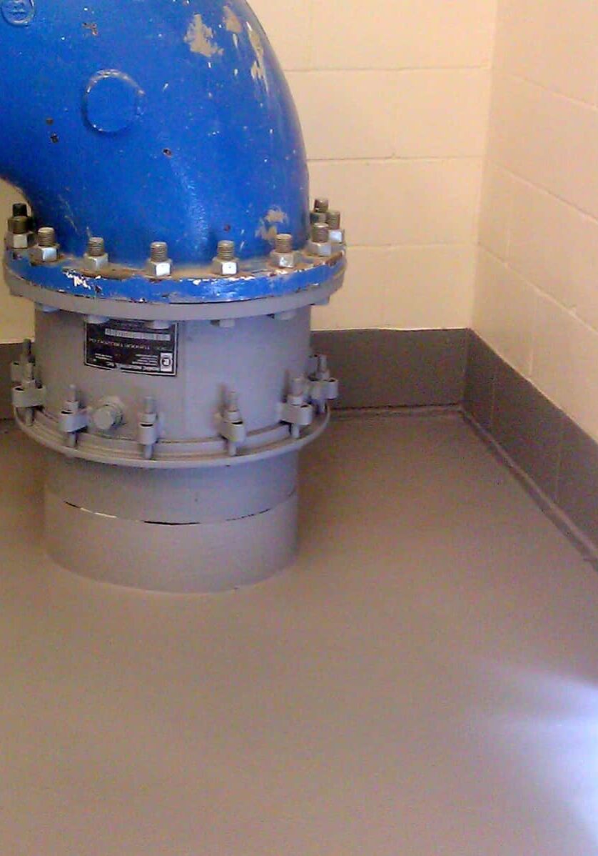 Large blue industrial pipe with a bolted joint emerges from the wall and extends into the floor of a room featuring white-painted brick walls and a gray surface finished with industrial floor coatings in Auburn, WA.